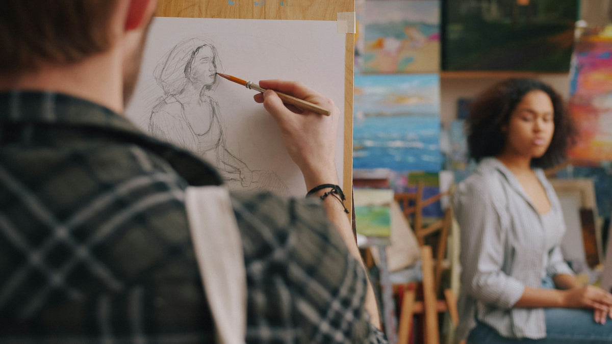Artist sketching a woman's portrait at an easel, with art canvases in the background
