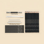 A flat-lay arrangement of an art supply set on a warm beige background, including a 12-pack of black graphite pencils with white text, a black and silver pencil extender, three white blending stumps, and a row of black pencil grips showing color options.