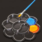 A flower-shaped clear plastic paint palette with multiple compartments, shown on a dark background with a blue-handled paintbrush resting in one section. Two paint wells contain blue and yellow paint.