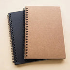 Two spiral-bound sketchpad on a light beige background, with one notebook in a brown kraft paper cover resting on top of another in a dark navy blue cover.