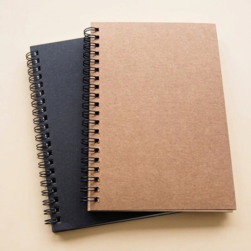 Two spiral-bound sketchpad on a light beige background, with one notebook in a brown kraft paper cover resting on top of another in a dark navy blue cover.